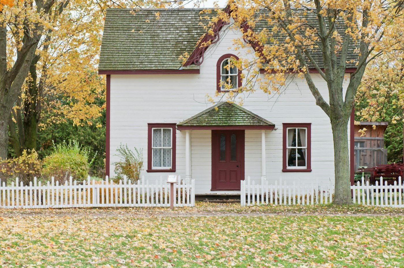 A white wooden house with a burgundy front door, a small porch, and a white picket fence, with autumn trees behind