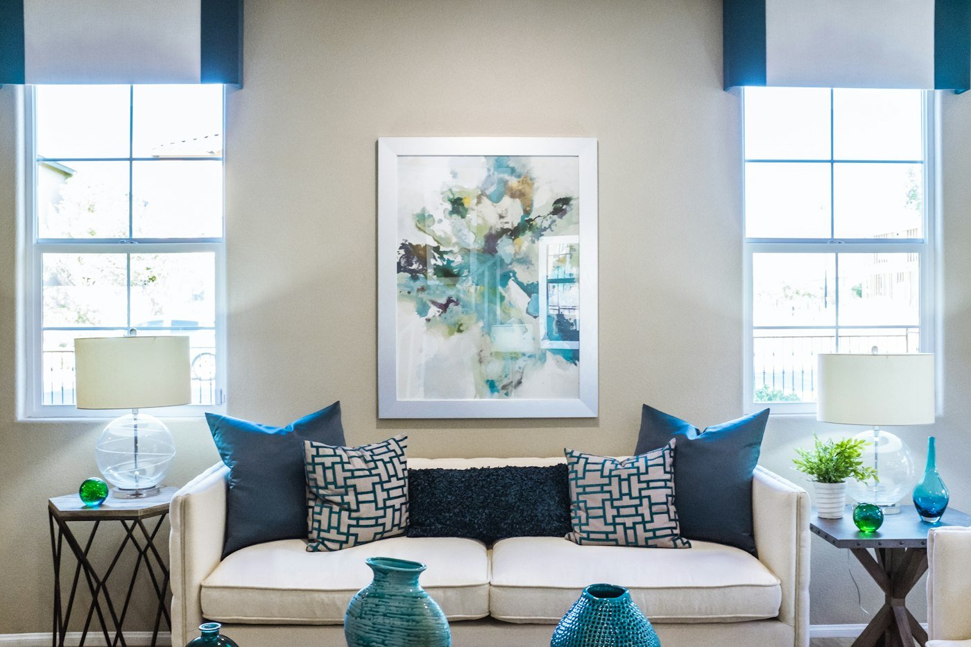 A cream sofa with dark blue and patterned cushions, between two windows with blue blinds